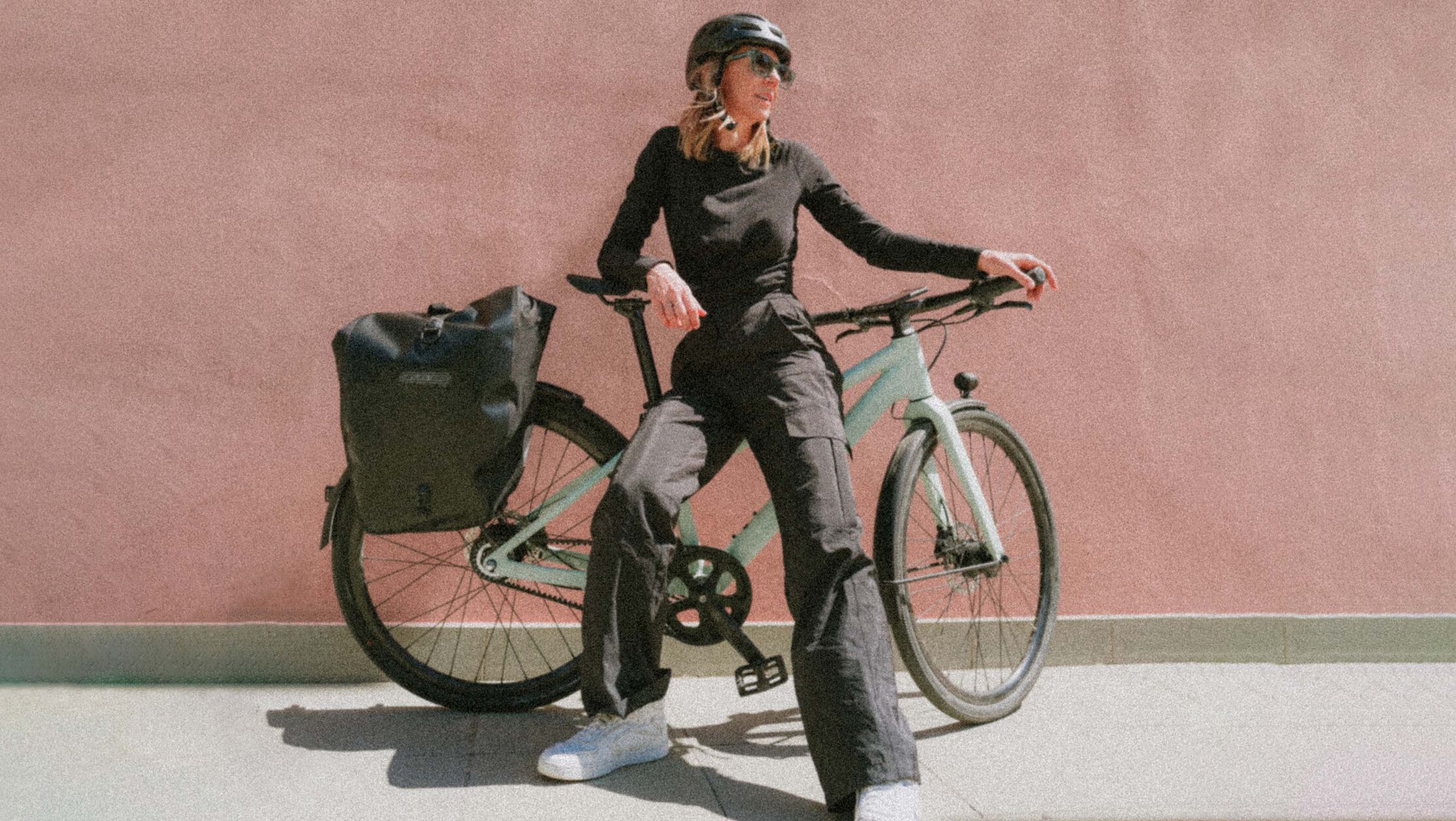 Woman posing with a Canyon commuter bike in front of a minimalist pink wall, wearing casual black outfit and helmet