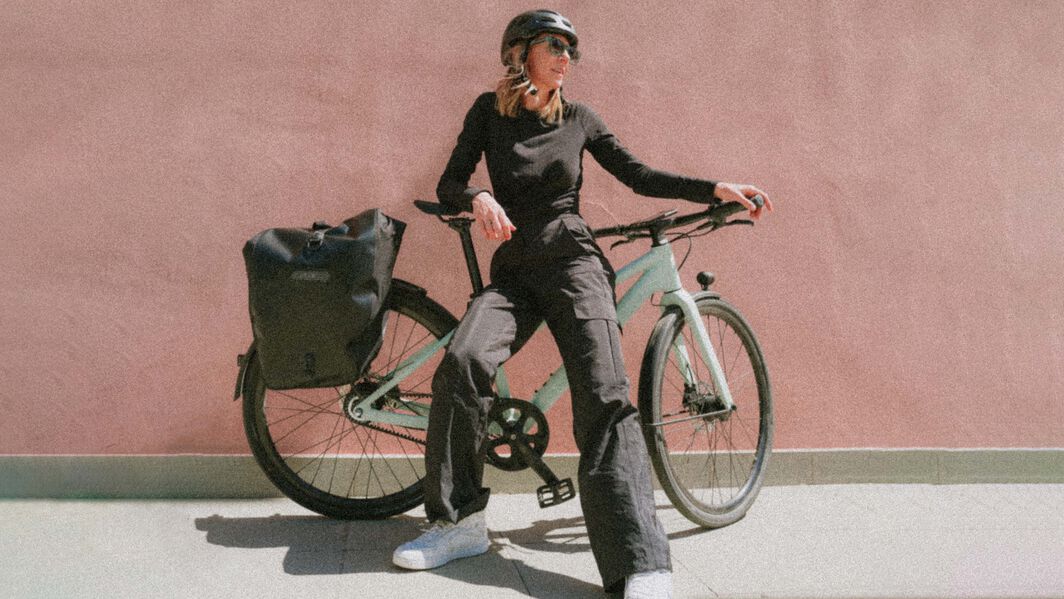 Woman posing with a Canyon commuter bike in front of a minimalist pink wall, wearing casual black outfit and helmet